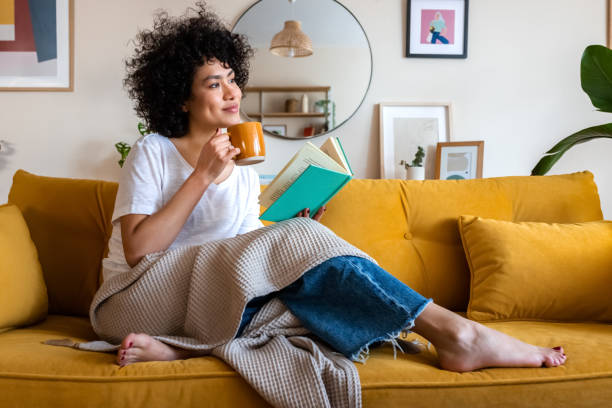 Relaxed woman sitting on a yellow couch, enjoying a cup of coffee while reading a book in a cozy living room