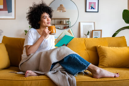 Relaxed woman sitting on a yellow couch, enjoying a cup of coffee while reading a book in a cozy living room